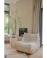 Ein gemütlicher, cremefarbener Struktursessel steht neben der künstlichen Pflanze Enkianthus 210 cm und einem modernen Kamin in einem hellen, minimalistischen Wohnzimmer mit hellen Holzböden und großen Fenstern.