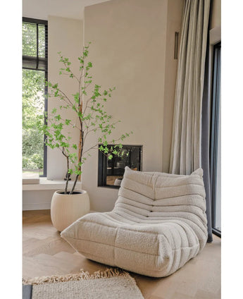 Ein gemütlicher, cremefarbener Struktursessel steht neben der künstlichen Pflanze Enkianthus 210 cm und einem modernen Kamin in einem hellen, minimalistischen Wohnzimmer mit hellen Holzböden und großen Fenstern.