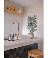 Eine moderne Arbeitsplatte verfügt über einen goldenen Wasserhahn, drei Seifenspender, die künstliche Eukalyptuspflanze (91 cm) und Holzhocker. Minimalistische Regale über den Gläsern mit verschiedenen Zutaten sorgen für eine gemütliche Atmosphäre.