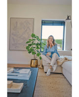 Eine Frau sitzt auf einem beigen Sofa in einem gemütlichen, modernen Wohnzimmer, hält eine Tasse in der Hand und lächelt. Der Raum wird durch die künstliche Ficus-Pflanze 120 cm in einem goldenen Topf, abstrakte Kunstwerke und Sonnenlicht, das durch blaue Jalousien scheint, geprägt.