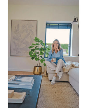 Eine Frau sitzt auf einem beigen Sofa in einem gemütlichen, modernen Wohnzimmer, hält eine Tasse in der Hand und lächelt. Der Raum wird durch die künstliche Ficus-Pflanze 120 cm in einem goldenen Topf, abstrakte Kunstwerke und Sonnenlicht, das durch blaue Jalousien scheint, geprägt.