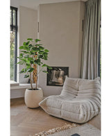 Ein gemütlicher, beiger Loungesessel mit Struktur steht neben der künstlichen Ficus-Pflanze 150 cm in einem modernen Wohnzimmer mit hellen Holzböden, neutralen Wänden, einem Fenster und bodenlangen grauen Vorhängen.