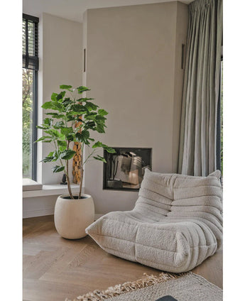 Ein gemütlicher, beiger Loungesessel mit Struktur steht neben der künstlichen Ficus-Pflanze 150 cm in einem modernen Wohnzimmer mit hellen Holzböden, neutralen Wänden, einem Fenster und bodenlangen grauen Vorhängen.