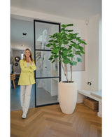 Eine Frau in gelbem Blazer und weißer Hose lächelt neben der Kunstpflanze Ficus 180 cm in einem modernen, hellen Raum mit Holzboden und minimalistischem Dekor.