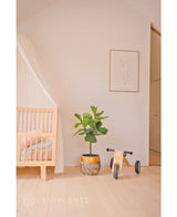 Ein minimalistisches Kinderzimmer mit einem hellen Holzbettgestell, einem durchscheinenden Baldachin, einer künstlichen Ficus-Tabakpflanze Deluxe 90 cm, Holzspielzeug mit der Aufschrift „Mia“ und einem minimalistischen Line-Art-gerahmten Druck auf einer schlichten weißen Wand.
