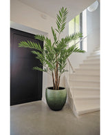 Die Kunstpflanze Hawaii Parlour 180 cm, eine hohe künstliche Palme für den Innenbereich mit üppigen grünen Blättern, steht auf einem grauen Boden in einer modernen Halle in der Nähe einer weißen Treppe und einer schwarzen Tür.