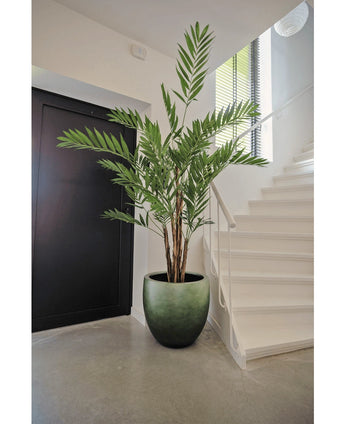 Die Kunstpflanze Hawaii Parlour 180 cm, eine hohe künstliche Palme für den Innenbereich mit üppigen grünen Blättern, steht auf einem grauen Boden in einer modernen Halle in der Nähe einer weißen Treppe und einer schwarzen Tür.