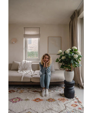 Eine Frau mit blonden Locken sitzt auf einem beigen Sofa in einem hellen Wohnzimmer und stützt ihr Kinn auf ihre Hände. Neben ihr steht die künstliche Hortensie UV-Pflanze, 85 cm hoch, beleuchtet durch ein Fenster mit beigen Vorhängen.