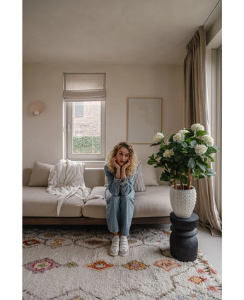 Eine Frau mit blonden Locken sitzt auf einem beigen Sofa in einem hellen Wohnzimmer und stützt ihr Kinn auf ihre Hände. Neben ihr steht die künstliche Hortensie UV-Pflanze, 85 cm hoch, beleuchtet durch ein Fenster mit beigen Vorhängen.