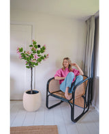 Eine Frau in rosa Pullover und blauen Jeans sitzt barfuß auf einem Rattanstuhl neben der Kunstpflanze Judas 150 cm, einem künstlichen Baum mit rosa Blüten, in einem hellen, modernen Raum mit weißen Wänden und Böden.