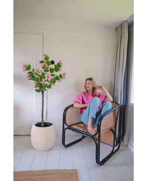 Eine Frau in rosa Pullover und blauen Jeans sitzt barfuß auf einem Rattanstuhl neben der Kunstpflanze Judas 150 cm, einem künstlichen Baum mit rosa Blüten, in einem hellen, modernen Raum mit weißen Wänden und Böden.