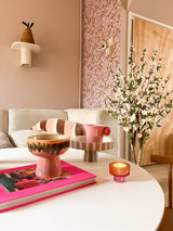 In einem gemütlichen Wohnzimmer steht ein weißer Couchtisch mit rosa Buch, Schale, Sockelschale, Kerze und der künstlichen Kirschblütenpflanze 120 cm creme. Im Hintergrund steht ein beiges Sofa mit gestreiften Kissen und rosa gemusterter Tapete.