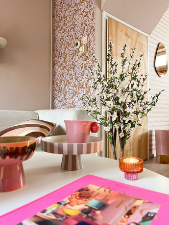 Ein gemütliches Wohnzimmer mit rosa und pastellfarbenem Dekor verfügt über ein gestreiftes Tablett auf dem Fuß, eine rosa Tasse, eine Kerze, eine Schale, eine lebhafte Zeitschrift und die künstliche Kirschblütenpflanze 120 cm cremefarben in einer hohen Vase neben einer gemusterten Wand und einer Holztür.