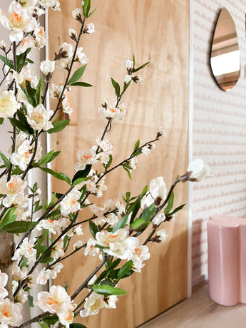 Vor einer Holztür steht eine künstliche Kirschblütenpflanze 120 cm creme mit weißen und hellrosa Blüten. Dahinter stehen ein rosa Hocker, ein Spiegel und eine rosa gemusterte Tapete.
