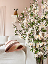 Ein gemütliches Wohnzimmer mit einem weißen Sofa, einem gestreiften rosa-braunen Kissen und dem 120 cm großen cremefarbenen Blütenzweig der künstlichen Kirschblüte im Vordergrund. Eine Topfpflanze steht auf einem Wandregal vor einer blassrosa Wand.
