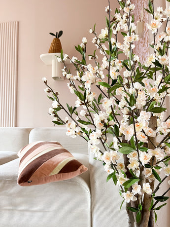 Ein gemütliches Wohnzimmer mit einem weißen Sofa, einem gestreiften rosa-braunen Kissen und dem 120 cm großen cremefarbenen Blütenzweig der künstlichen Kirschblüte im Vordergrund. Eine Topfpflanze steht auf einem Wandregal vor einer blassrosa Wand.