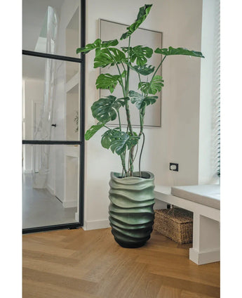 Die Kunstpflanze Monstera 150 cm mit ihren großen grünen Blättern steht in einem Topf mit Wellenmuster auf einem Holzboden in einem modernen, hellen Raum mit Glastür, gerahmter Kunst und einem Weidenkorb daneben.
