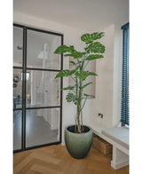 Die Kunstpflanze Monstera 180 cm, eine hohe Topfpflanze mit großen gespaltenen Blättern, steht in einem modernen, hellen Raum neben einer Glastür, Holzboden, Sofa und blauen vertikalen Lamellen.