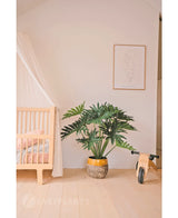 Ein gemütliches, minimalistisches Kinderzimmer mit einer hölzernen Wiege und einem Baldachin auf der linken Seite, einer künstlichen Philodendron UV 90 cm Pflanze in der Mitte als Prunkstück, einem hölzernen Spielzeugfahrrad auf der rechten Seite und einem einfachen Line-Art-Druck darüber.