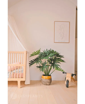 Ein gemütliches, minimalistisches Kinderzimmer mit einer hölzernen Wiege und einem Baldachin auf der linken Seite, einer künstlichen Philodendron UV 90 cm Pflanze in der Mitte als Prunkstück, einem hölzernen Spielzeugfahrrad auf der rechten Seite und einem einfachen Line-Art-Druck darüber.