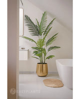 Die Kunstpflanze Phönix 180 cm in einem goldenen Topf steht neben einer weißen Badewanne und einem Waschbecken in einem modernen, minimalistischen Badezimmer mit weißen Wänden und Boden. Daneben liegt eine runde, gewebte Matte auf dem Boden.