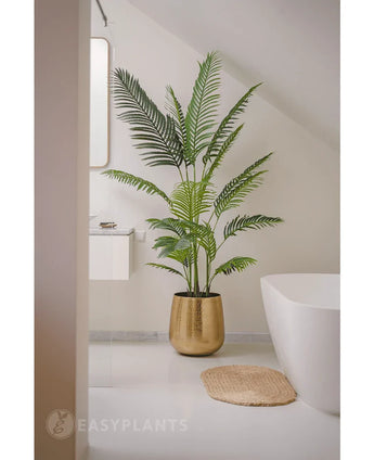 Die Kunstpflanze Phönix 180 cm in einem goldenen Topf steht neben einer weißen Badewanne und einem Waschbecken in einem modernen, minimalistischen Badezimmer mit weißen Wänden und Boden. Daneben liegt eine runde, gewebte Matte auf dem Boden.