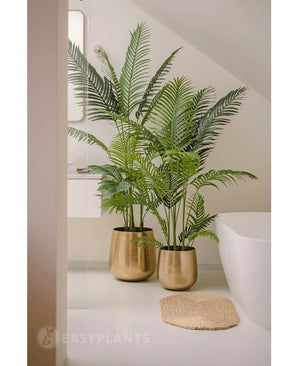 Eine 180 cm große Kunstpflanze Phönix mit sattgrünen Blättern in einem goldglänzenden Topf steht neben einer weißen Badewanne und einer beigen gewebten Matte in einem modernen, minimalistischen Badezimmer, ergänzt durch einen Spiegel und ein Fenster für einen ruhigen Look.