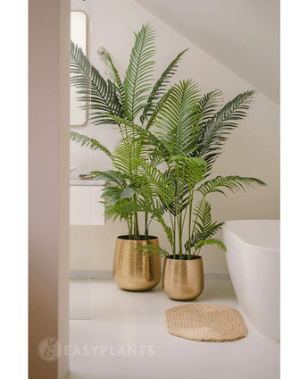 Eine 180 cm große Kunstpflanze Phönix mit sattgrünen Blättern in einem goldglänzenden Topf steht neben einer weißen Badewanne und einer beigen gewebten Matte in einem modernen, minimalistischen Badezimmer, ergänzt durch einen Spiegel und ein Fenster für einen ruhigen Look.