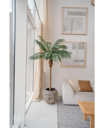 De Kunstplant Phoenix Palm De Luxe 180 cm in een pot met structuur staat bij grote ramen in een lichte woonkamer met neutrale inrichting, met een witte bank, beige kussens, een houten tafel en abstracte kunst aan de muur.
