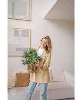 Eine Frau in einem beigen Blazer und Jeans steht in einem modernen Wohnzimmer und lächelt, während sie eine künstliche Pflanze Podocarpus 47 cm in der Hand hält. Abstrakte Kunst schmückt die helle Wand über einem cremefarbenen Sofa mit braunen Kissen.