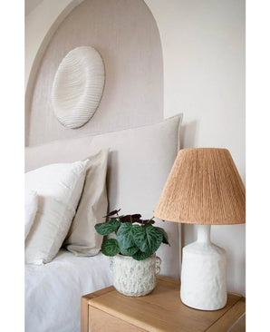 Ein modernes Schlafzimmer mit einer neutralen Farbpalette verfügt über ein Bett mit weißer Bettwäsche, einen Nachttisch aus Holz, eine strukturierte Keramiklampe mit gewebtem Schirm, die künstliche Saxifraga-Pflanze 27 cm und eine runde weiße Wandskulptur über dem Kopfteil.