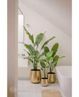Eine künstliche Strelitzia Nicolai Deluxe 120 cm Pflanze steht in einem glänzenden goldenen Pflanzgefäß auf dem Boden eines modernen, minimalistischen Badezimmers mit einer weißen Badewanne und hellen Wänden.