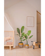 Ein gemütliches Kinderzimmer mit einem hölzernen Kinderbett, einer luxuriösen Kunstpflanze Strelitzia Nicolai 120 cm, einer gerahmten minimalistischen Strichzeichnung an der Wand und einem kleinen hölzernen Laufrad mit der Aufschrift MIA auf einem hellen Holzboden.