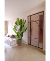 Ein modernes Wohnzimmer mit einem weißen Sofa, gemusterten Kissen und der Kunstpflanze Strelitzia Real Touch Deluxe 210 cm. Ein heller Holzboden, zartrosa Wände und eine Glastür mit bronzefarbenem Metallrahmen sorgen für eine helle, luftige Atmosphäre.