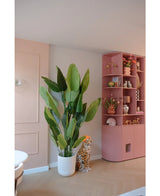 Die Kunstpflanze Strelitzia Real Touch Deluxe 210 cm steht neben einer Gepardenfigur aus Keramik mit orangefarbenem Hut, neben einem hohen rosa Regal voller Vasen, Blumen und Ornamenten in einem pastellfarbenen Raum.