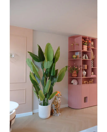 Die Kunstpflanze Strelitzia Real Touch Deluxe 210 cm steht neben einer Gepardenfigur aus Keramik mit orangefarbenem Hut, neben einem hohen rosa Regal voller Vasen, Blumen und Ornamenten in einem pastellfarbenen Raum.