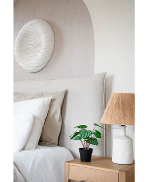 Ein gemütliches Schlafzimmer verfügt über ein Bett mit neutraler Bettwäsche, einen hellen Nachttisch aus Holz mit der künstlichen Wassermelonenpflanze 29 cm und eine weiße Lampe mit Korbschirm. Über dem Bett hängt eine weiß strukturierte Wandskulptur.