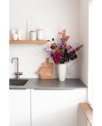 Eine moderne weiße Küche mit einer grauen Arbeitsplatte, einer Spüle, einem Holzschneidebrett und einer weißen Vase gefüllt mit einer künstlichen Hortensie 55 cm in Dunkelrosa. Weiße Tassen und Töpfe stehen auf einem Holzregal über der Theke.