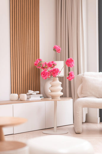 Ein modernes Wohnzimmer verfügt über einen weißen Beistelltisch mit dem künstlichen Zweig Kirschblüte 85 cm rosa, zu dem leuchtend rosa Blüten hinzugefügt wurden. Neutrales Dekor, helle Holzakzente, beige Vorhänge und ein strukturierter Sessel runden den gemütlichen, minimalistischen Raum ab.