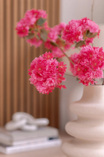 Eine weiße Keramikvase enthält einen künstlichen Kirschblütenzweig 85 cm rosa. Der Hintergrund zeigt vertikale Holztafeln, einen Stapel Bücher und darauf ein kleines Dekorationsobjekt.