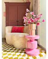 Ein gemütliches Wohnzimmer mit einem beigen runden Sofa, einem rosa und orangefarbenen geometrischen Kissen, einem rosa Beistelltisch, einem künstlichen Magnolienzweig mit kleinen rosa Blüten (89 cm) in einer Vase, einem gelb-weiß gemusterten Teppich und einem kastanienbraunen Schrank im Hintergrund.