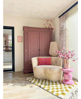 Eine gemütliche Leseecke verfügt über einen cremefarbenen Stuhl, rosa Kissen, einen gelb gemusterten Teppich und einen malvenfarbenen Schrank. Der künstliche Zweig Magnolia kleinblütig 89 cm rosa steht auf einem rosa Beistelltisch, während große Fenster diesen einladenden Raum mit sanftem Licht füllen.