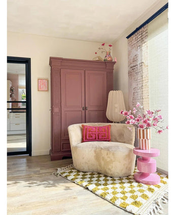 Eine gemütliche Leseecke verfügt über einen cremefarbenen Stuhl, rosa Kissen, einen gelb gemusterten Teppich und einen malvenfarbenen Schrank. Der künstliche Zweig Magnolia kleinblütig 89 cm rosa steht auf einem rosa Beistelltisch, während große Fenster diesen einladenden Raum mit sanftem Licht füllen.