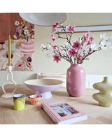 Ein moderner Raum verfügt über einen gedeckten Tisch mit künstlichem Magnolienzweig, kleinblumig, 89 cm groß, rosa in einer rosa Vase, pastellfarbenen Kerzenständern, Schalen, Tassen, einer grünen Skulpturenvase, einem rosa Buch und Blumenkunstwerken an der Wand.