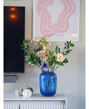 Auf einem weißen Schrank steht eine blaue Glasvase mit einer künstlichen Zweigbeere (76 cm) in Lila. Daneben stehen zwei weiße Kürbisse mit goldenen Stielen. An der Wand dahinter hängen ein Fernseher und ein abstrakter rosa Kunstdruck.
