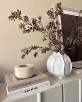 Auf einem hellen Schrank steht eine weiße Vase, gefüllt mit der künstlichen Zweig-Eukalyptus-Beere 110 cm braun, daneben ein Stapel Bücher und ein Steinkerzenständer, im Hintergrund ein gerahmtes Foto.