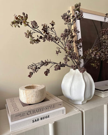 Auf einem hellen Schrank steht eine weiße Vase, gefüllt mit der künstlichen Zweig-Eukalyptus-Beere 110 cm braun, daneben ein Stapel Bücher und ein Steinkerzenständer, im Hintergrund ein gerahmtes Foto.