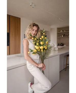 Eine Frau mit lockigen blonden Haaren, gekleidet in ein cremefarbenes Outfit, lehnt an einer Theke und hält den Blumenstrauß „Künstlicher Zitronenzweig 104 cm gelb“, dekoriert mit üppigem Grün. Die moderne Küche ist in neutralen Farben gehalten und verfügt über Holzakzente.