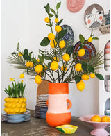 Ein heller Raum mit einer hohen Vase voller Zitronen und grünen Blättern, in der das auffällige Kunsttak Lemon 104 cm Gelb einen prominenten Platz im Arrangement einnimmt, umgeben von bunten Keramikschalen, einer Schale mit Zitronen und dekorativen Tellern an der Wand – so entsteht ein fröhlicher, vielseitiger Look.