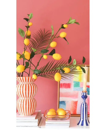 Vor einer korallenrosa Wand stehen eine gestreifte Vase mit Kunsttak-Zitronenzweigen von 104 cm in Gelb, ein farbenfrohes Gemälde, ein Stapel Bücher, ein blauer Kerzenständer mit einer Kerze und ein Tablett mit Zitronen.